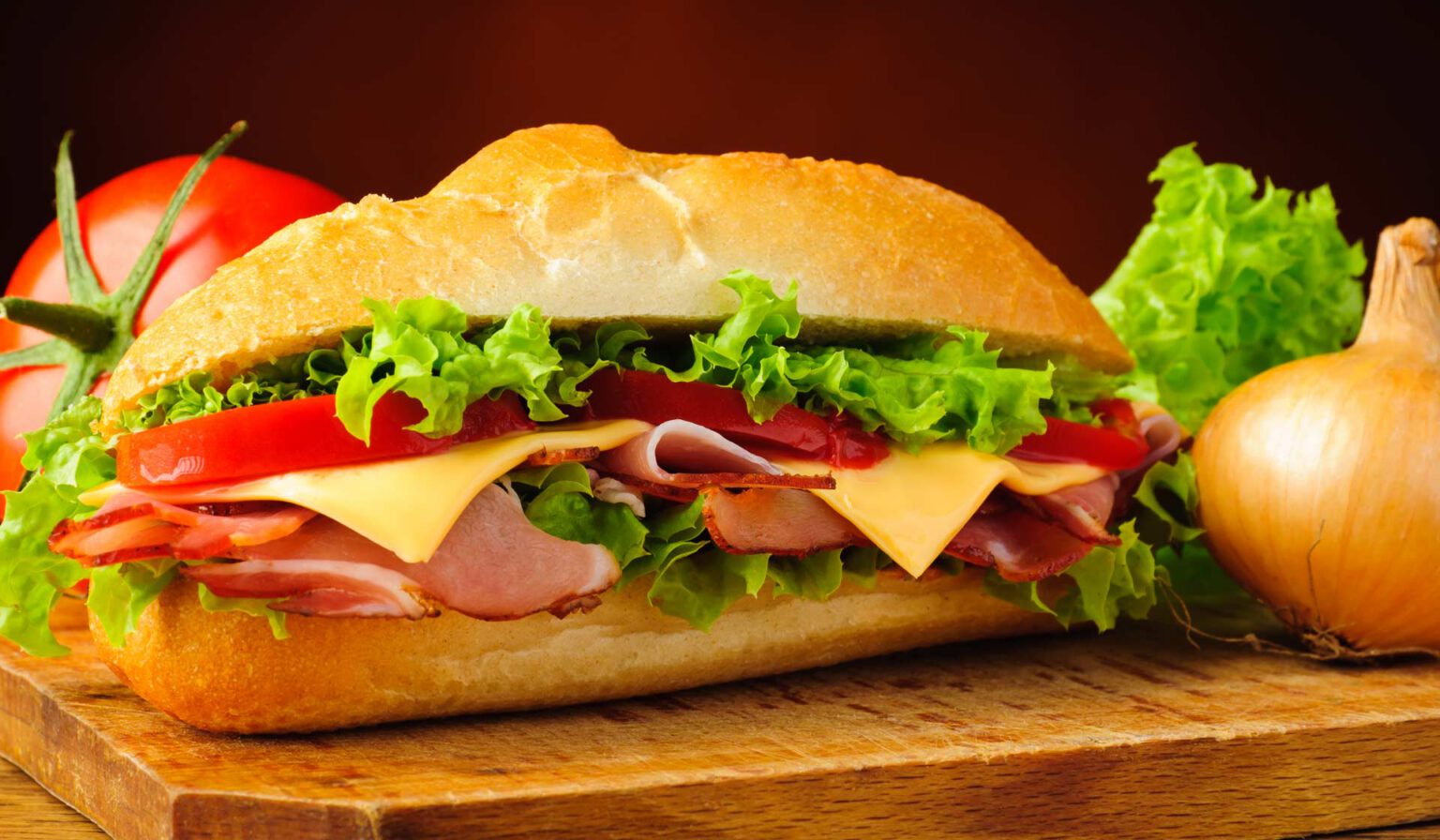 A loaded deli sandwich with lettuce, tomatoe, cheese, and meat. There is a tomato and omion in the background.