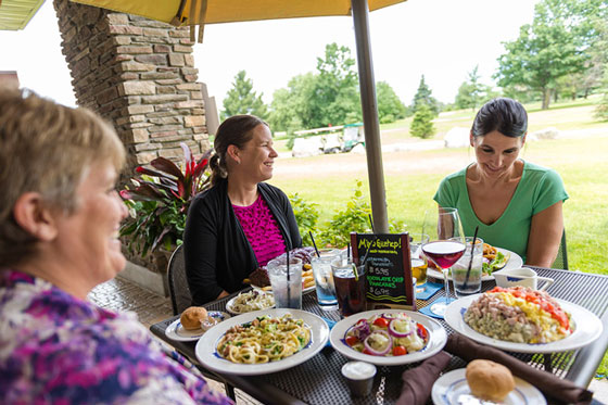 Three women sitting at a table outdoors under an umbrella, enjoying a meal. Plates of food, including pasta, salads, and other dishes, are on the table. The background features green trees and a lawn, indicating a pleasant setting.